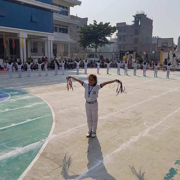 Children in white uniforms participate in a cultural event on an outdoor sports ground.